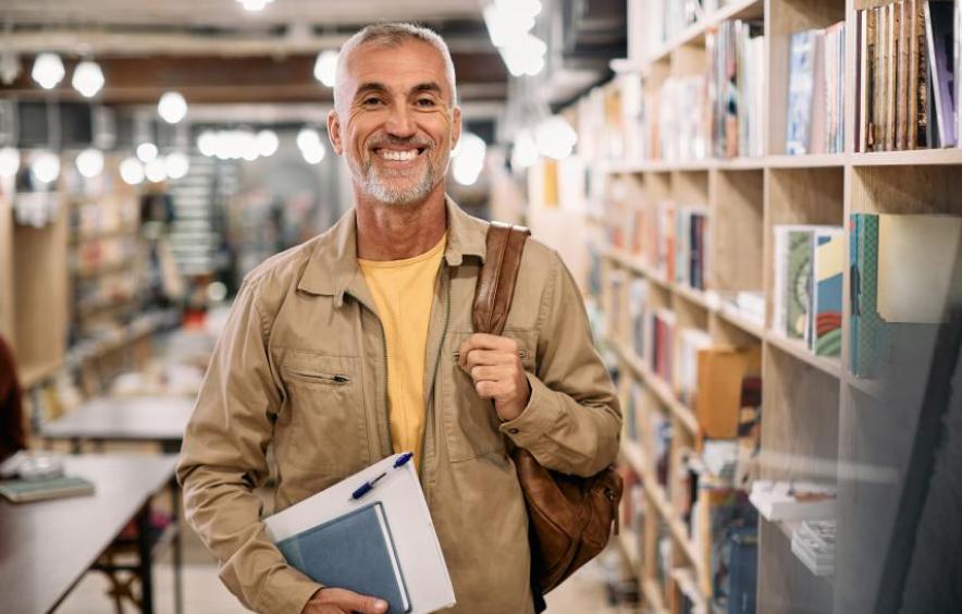 man in library with backpack on one shoulder and notebook in hand