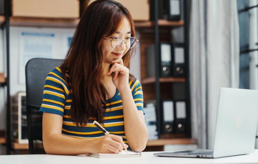 Asian woman with glasses and long dark hair, wearing a striped short sleeve shirt, sitting at laptop