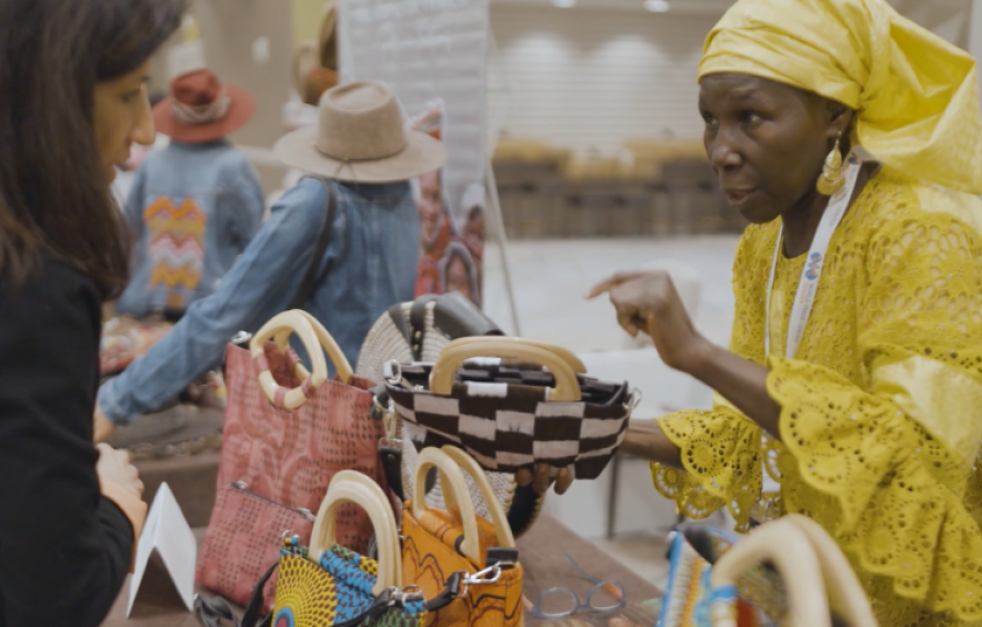 african woman in yellow outfit speaks to a customer about her handbags